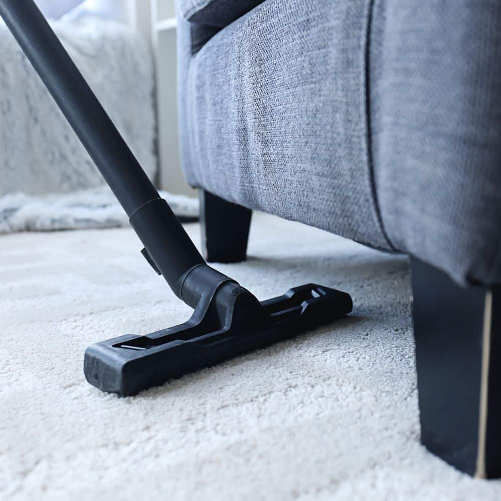 Chorda Carpet Cleaning Services in West Sussex | Close up image of the black wooden feet of a grey sofa, on a cream carpet which is being vacuumed by a black hoover.